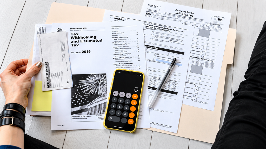 Person reviewing 2019 tax withholding and estimated tax documents including W-2, 1040-ES, and Publication 505 with smartphone calculator on white desk